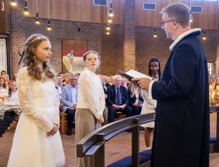 To piger i hvide konfirmationskjoler står foran præsten, der læser et konfirmationsord på for ene konfirmand. 