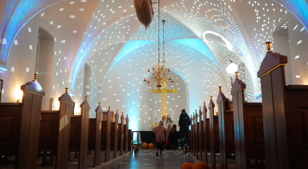 Kirkerummet i Lønstrup Kirke med græskar på gulvet, børn ved alteret og lys på vægge og lofter