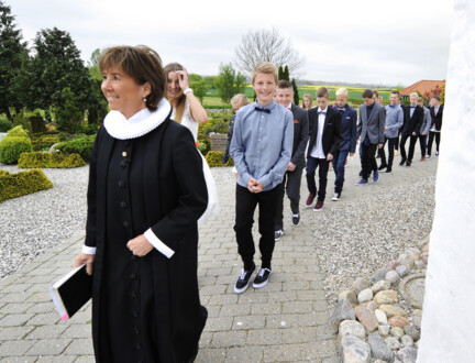 En præst går foran en lang række glade konfirmander uden for en kirke.