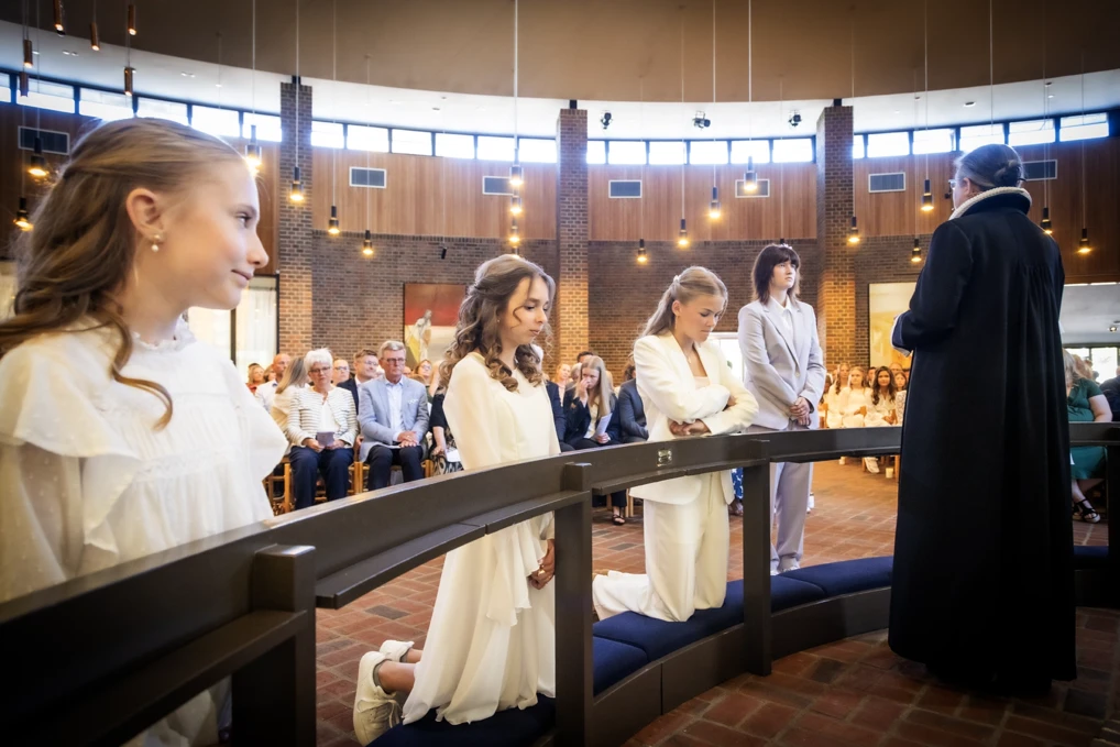 Konfirmander og præst 15 konfirmander er samlet foran alteret i en kirke, hvor præsten er i gang med at konfirmere en dreng ved at lægge hånden på hans hoved.